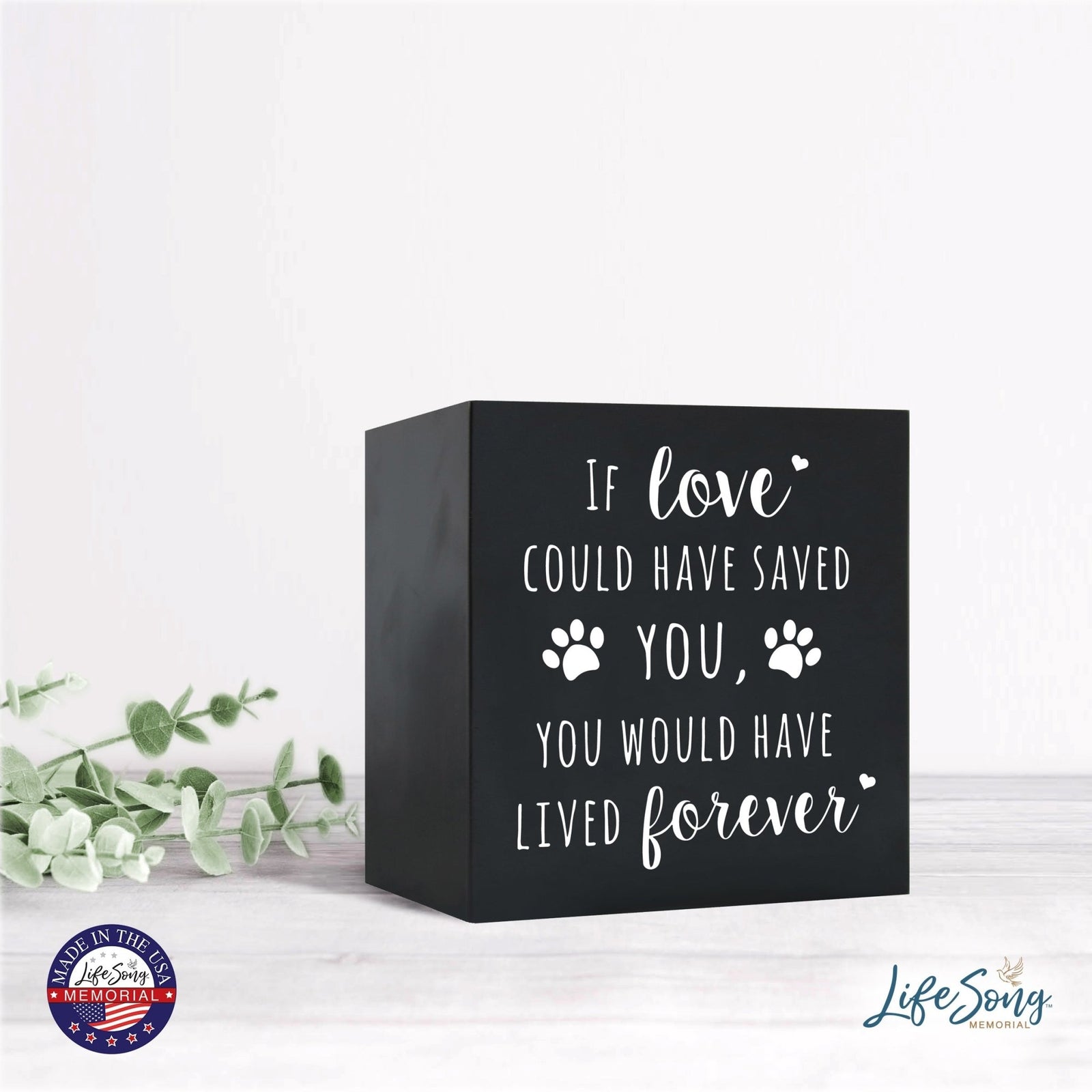 A black Lifesong Milestones Memorial Shadow Box Urn For Pet Ashes with an engraved message accompanied by two paw prints. The urn sits on a light-colored wooden surface, decorated with greenery, creating a serene and heartfelt tribute.