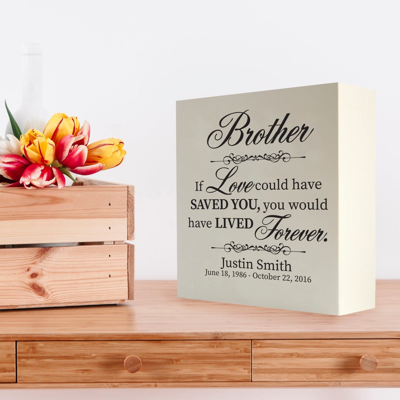 Custom white wooden shadow box urn for human ashes adult, inscribed with 'Brother: If love could have saved you, you would have lived forever,' placed on a wooden dresser adorned with vibrant tulips.