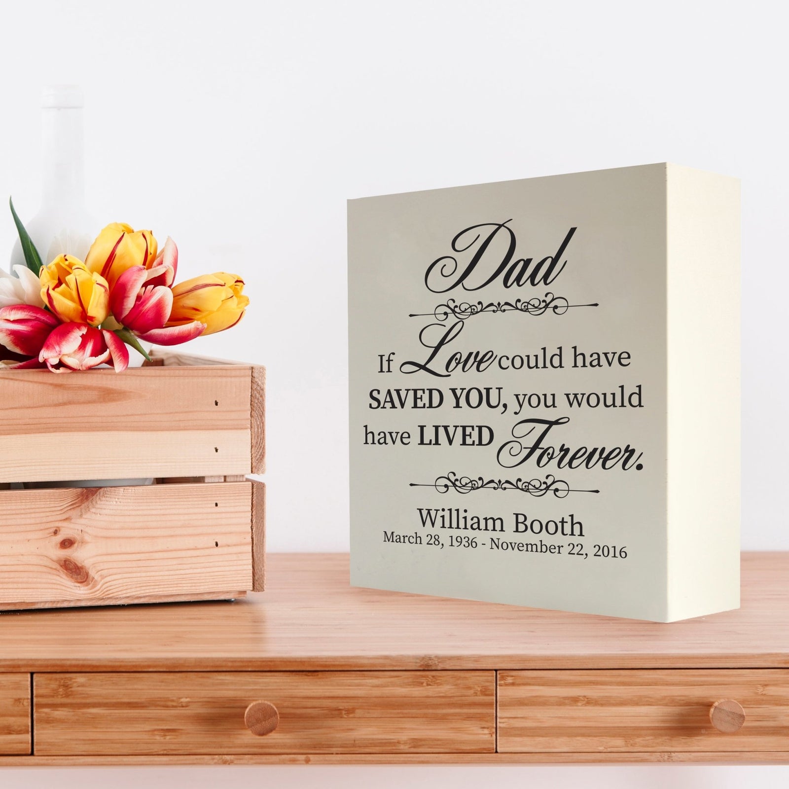 Custom white wooden shadow box urn for human ashes adult, inscribed with 'Dad: If love could have saved you, you would have lived forever,' placed on a wooden dresser adorned with vibrant tulips.