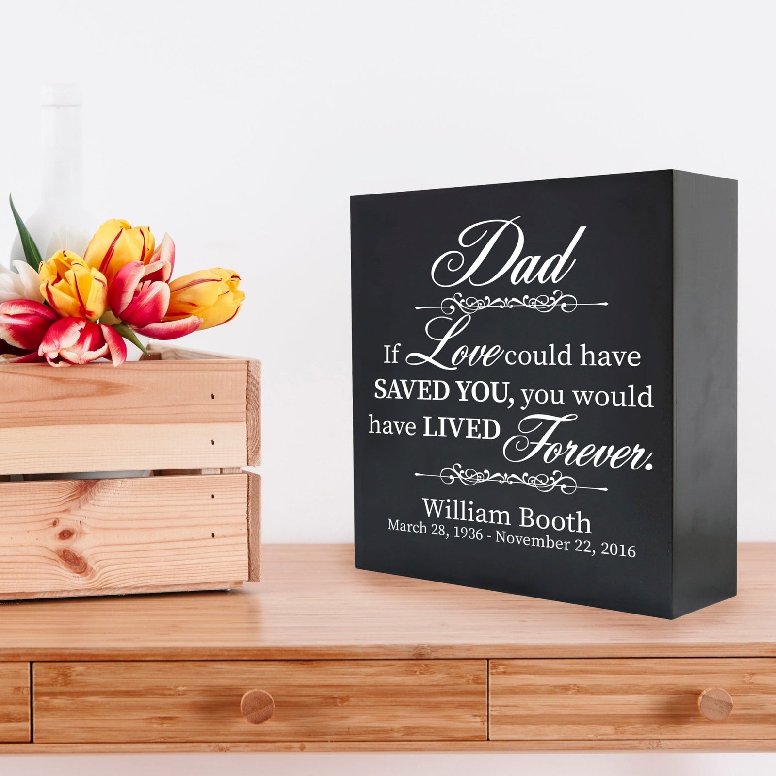 Custom black wooden shadow box urn for human ashes adult, inscribed with 'Dad: If love could have saved you, you would have lived forever,' placed on a wooden dresser adorned with vibrant tulips.