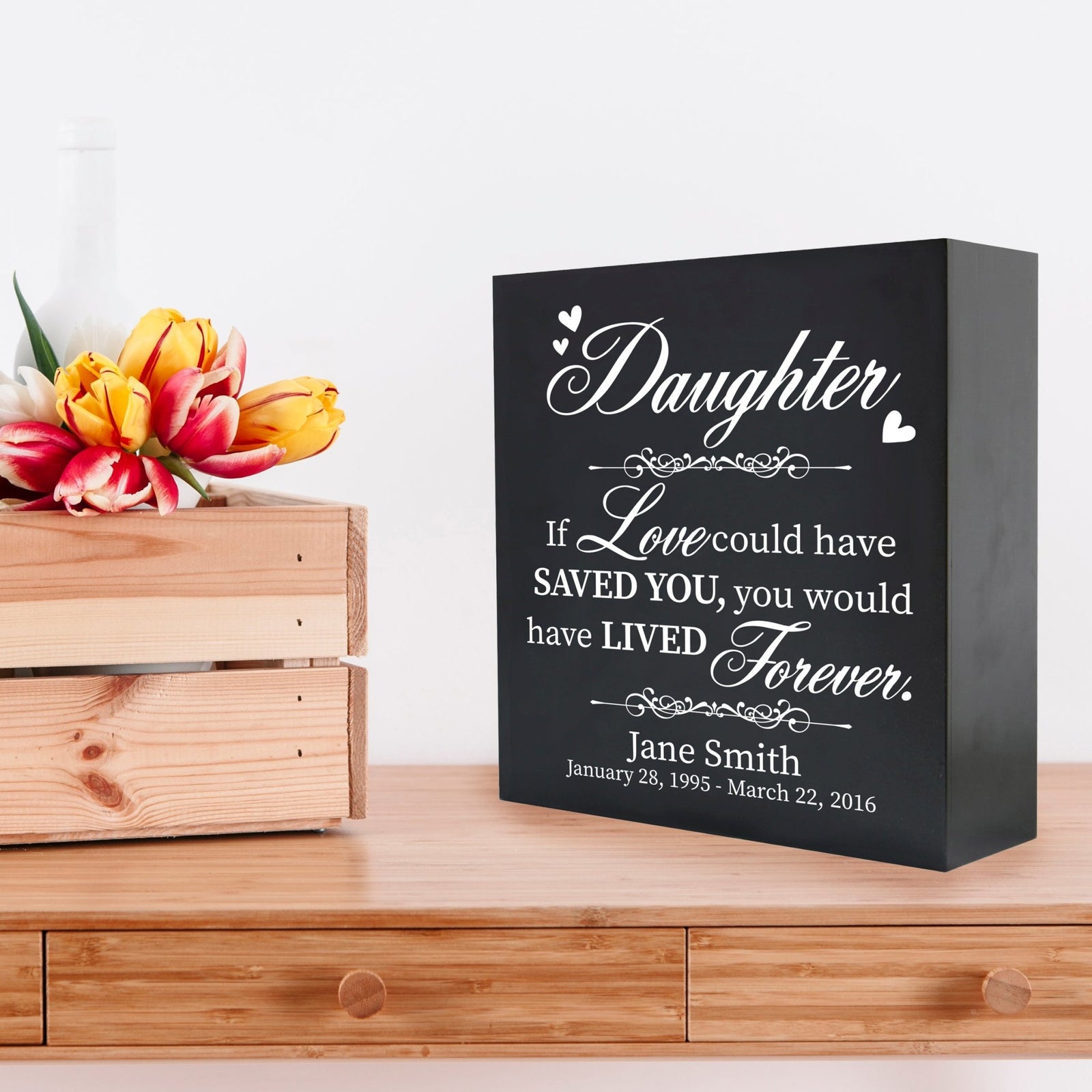 A black wooden shadow box urn for human ashes, designed for adults, featuring a heartfelt tribute with the text " daughter: If love could have saved you, you would have lived forever" in white lettering. The box sits elegantly on a wooden table next to a crate of vibrant tulips, creating a serene and comforting remembrance piece.