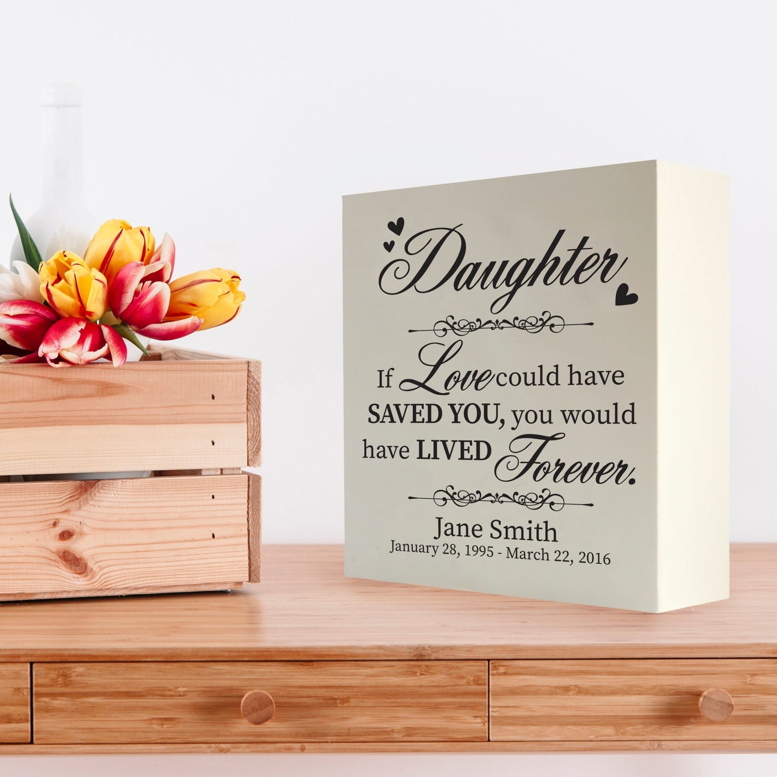 Custom white wooden shadow box urn for human ashes adult, inscribed with daughter: If love could have saved you, you would have lived forever,' placed on a wooden dresser adorned with vibrant tulips.