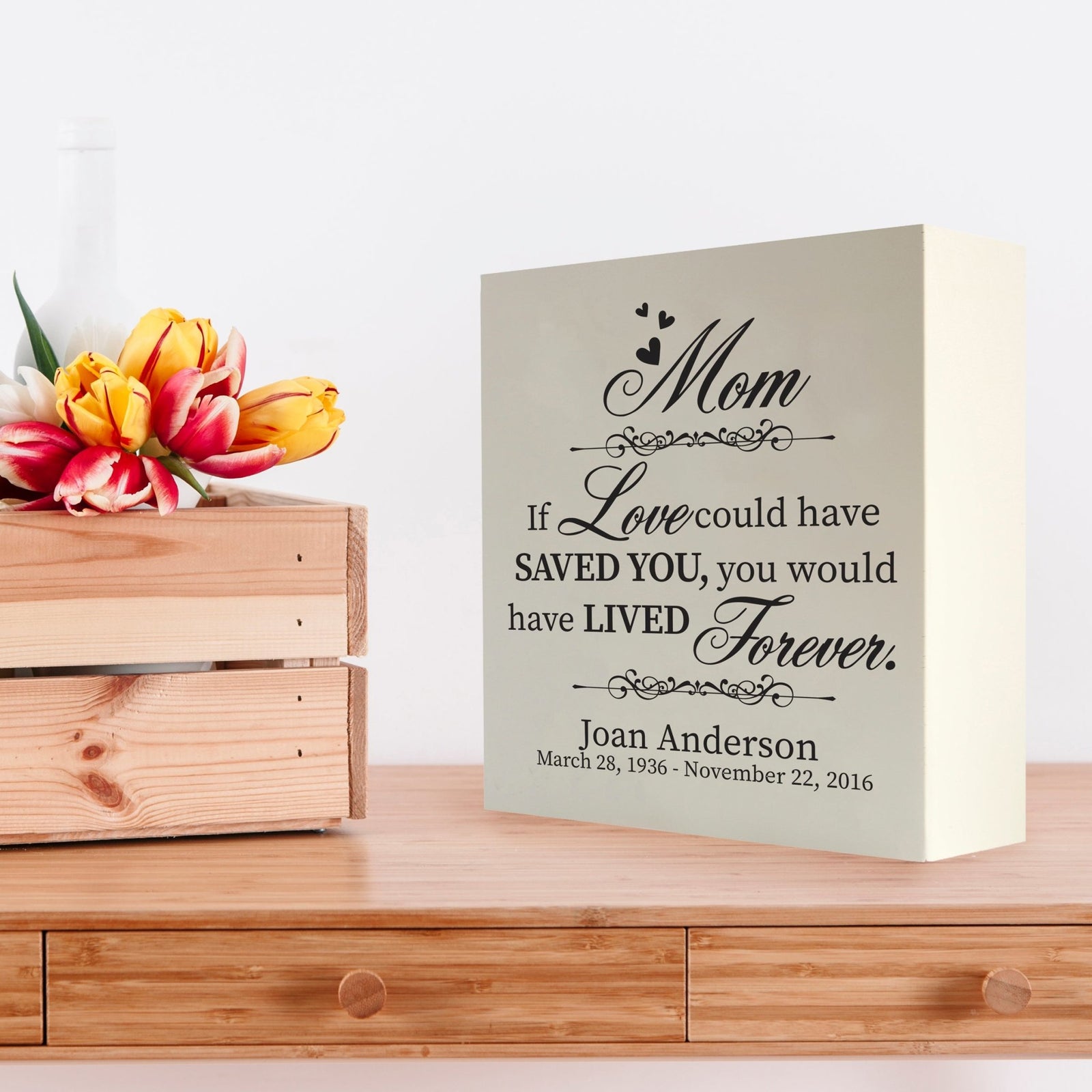 Custom white wooden shadow box urn for human ashes adult, inscribed with mom: If love could have saved you, you would have lived forever,' placed on a wooden dresser adorned with vibrant tulips.