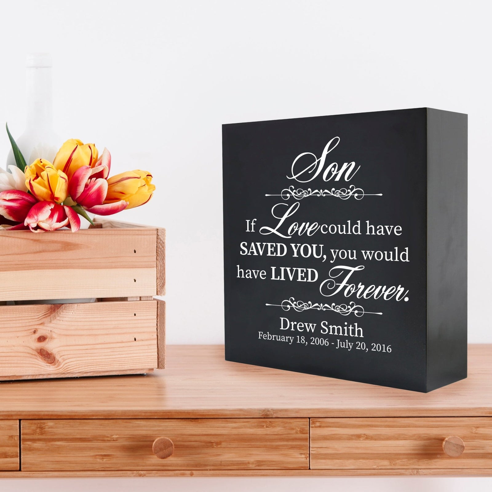 A black wooden shadow box urn for human ashes, designed for adults, featuring a heartfelt tribute with the text " son: If love could have saved you, you would have lived forever" in white lettering. The box sits elegantly on a wooden table next to a crate of vibrant tulips, creating a serene and comforting remembrance piece.