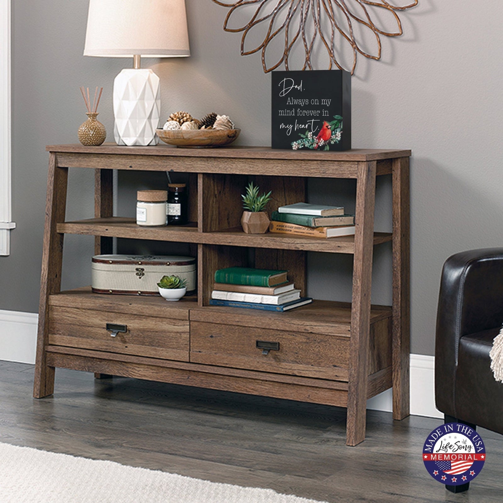 Rustic wooden console table with multiple shelves, decorated with a cream-colored wooden shadow box urn for human ashes. The urn features a red cardinal and floral accents. Surrounded by books, potted plants, and a textured white lamp, this handcrafted adult memorial urn blends seamlessly into a cozy living room setting. Perfect for those seeking wooden shadow box urns for human ashes, offering a beautiful and personalized tribute.