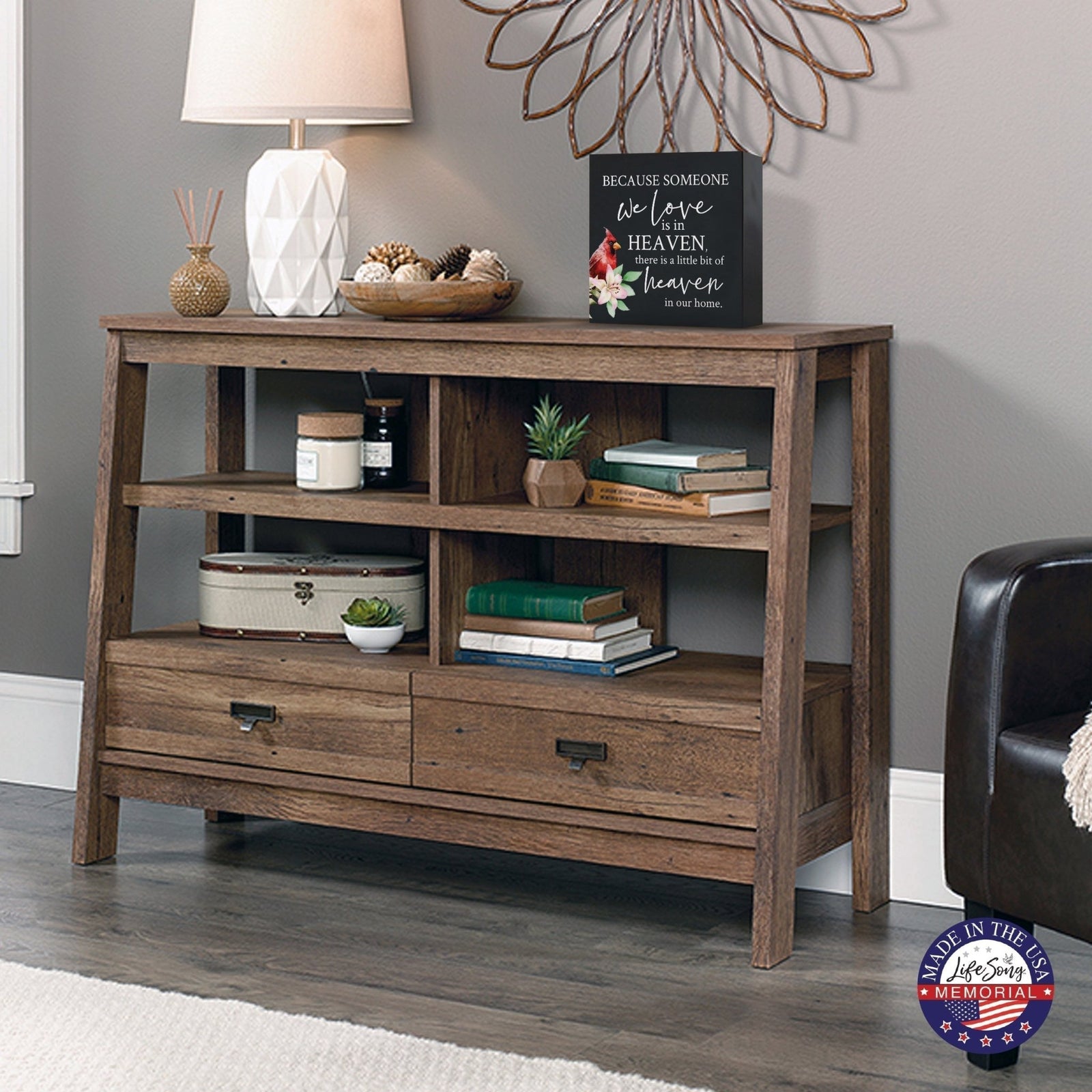 Rustic wooden console table with multiple shelves, decorated with a black-colored wooden shadow box urn for human ashes. The urn features a red cardinal and floral accents. Surrounded by books, potted plants, and a textured white lamp, this handcrafted adult memorial urn blends seamlessly into a cozy living room setting. Perfect for those seeking wooden shadow box urns for human ashes, offering a beautiful and personalized tribute.