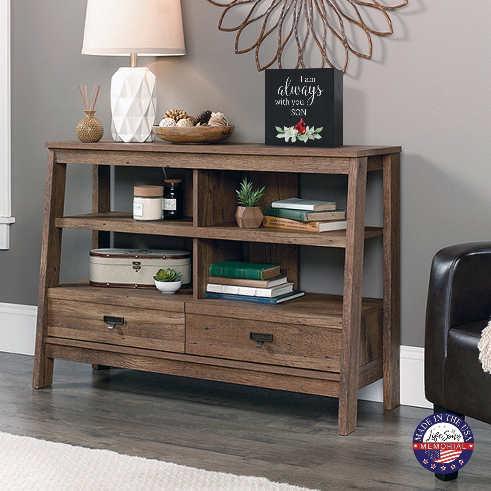 Rustic wooden console table with multiple shelves, decorated with a cream-colored wooden shadow box urn for human ashes. The urn features a red cardinal and floral accents. Surrounded by books, potted plants, and a textured white lamp, this handcrafted adult memorial urn blends seamlessly into a cozy living room setting. Perfect for those seeking wooden shadow box urns for human ashes, offering a beautiful and personalized tribute.