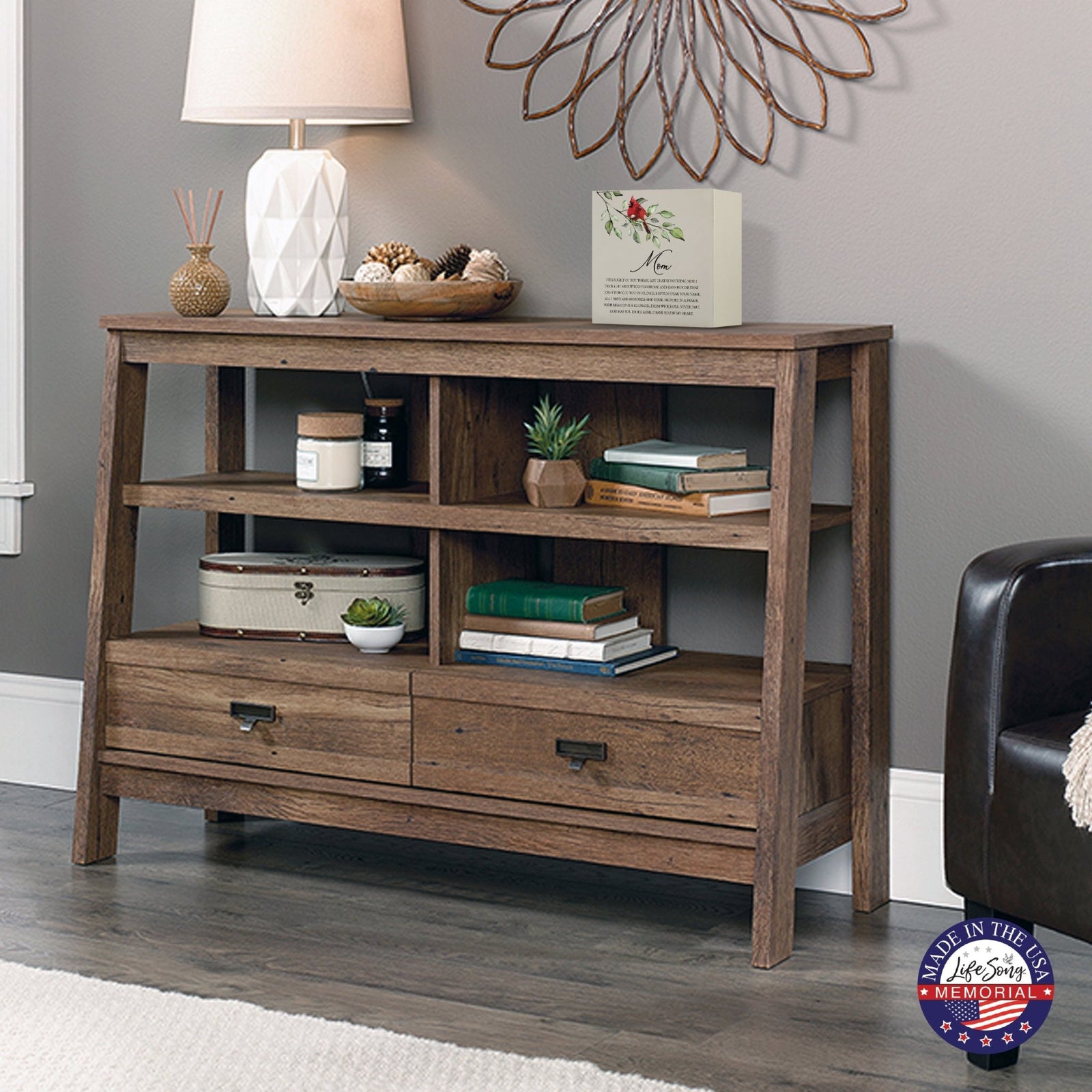 Rustic wooden console table with multiple shelves, decorated with a cream-colored wooden shadow box urn for human ashes. The urn features a red cardinal and floral accents. Surrounded by books, potted plants, and a textured white lamp, this handcrafted adult memorial urn blends seamlessly into a cozy living room setting. Perfect for those seeking wooden shadow box urns for human ashes, offering a beautiful and personalized