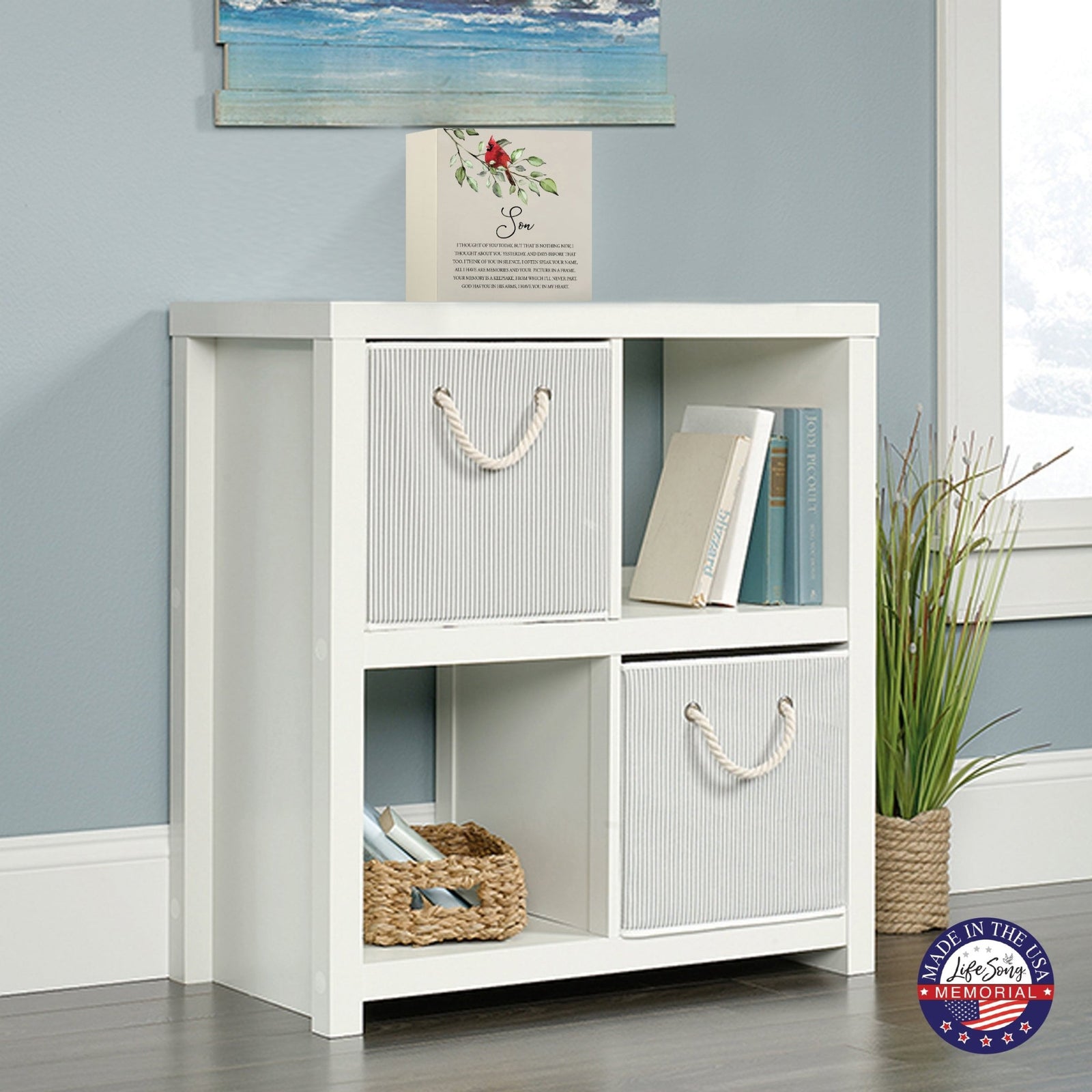 White wooden storage unit topped with a decorative wooden shadow box urn for son. The urn features a red cardinal illustration and floral accents. This adult memorial urn is displayed in a cozy living space with a beach-themed painting above.