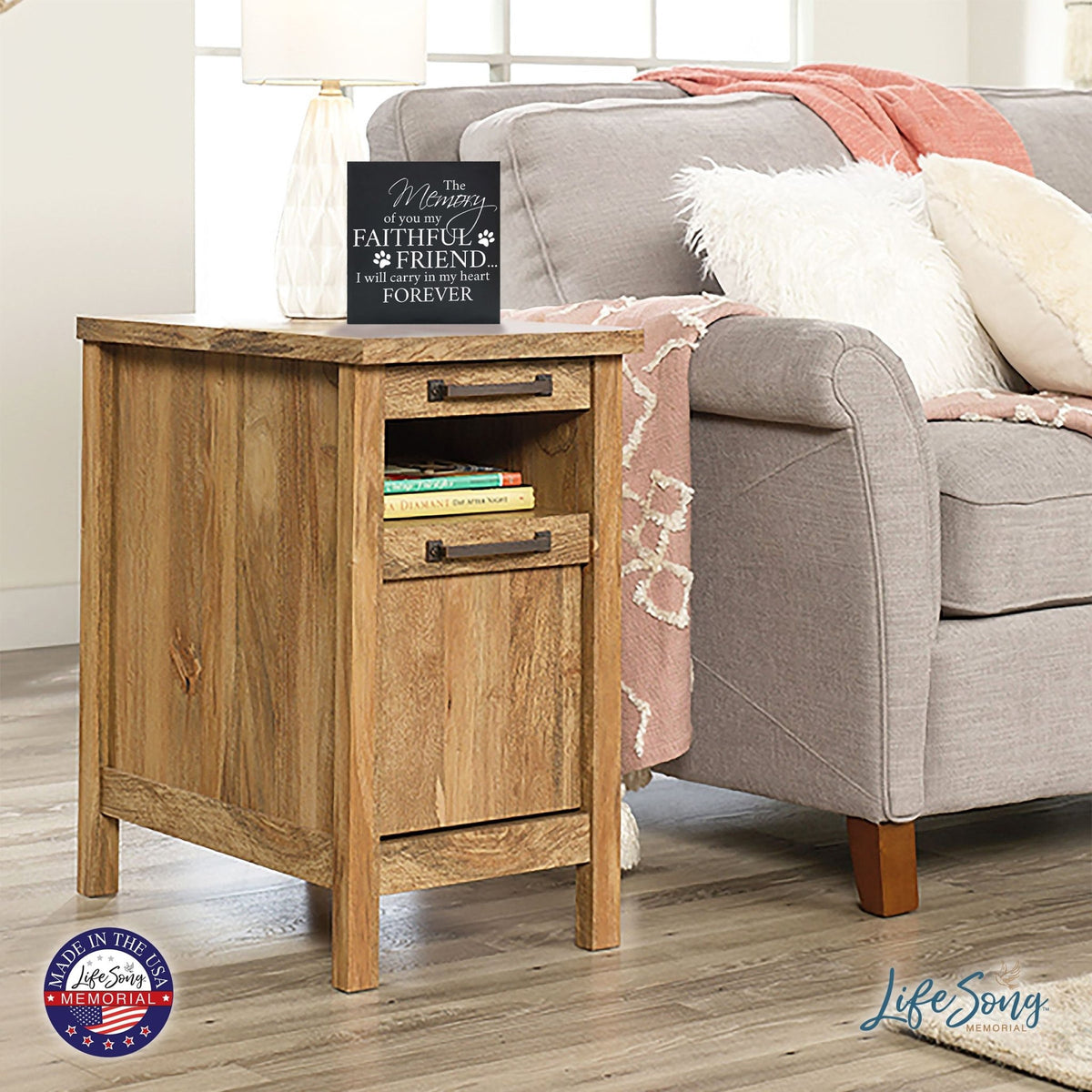 A black Lifesong Milestones Memorial Shadow Box Urn For Pet Ashes resting on a rustic wooden side table next to a cozy gray sofa with soft cushions. The urn features an emotional inscription, honoring a cherished pet.