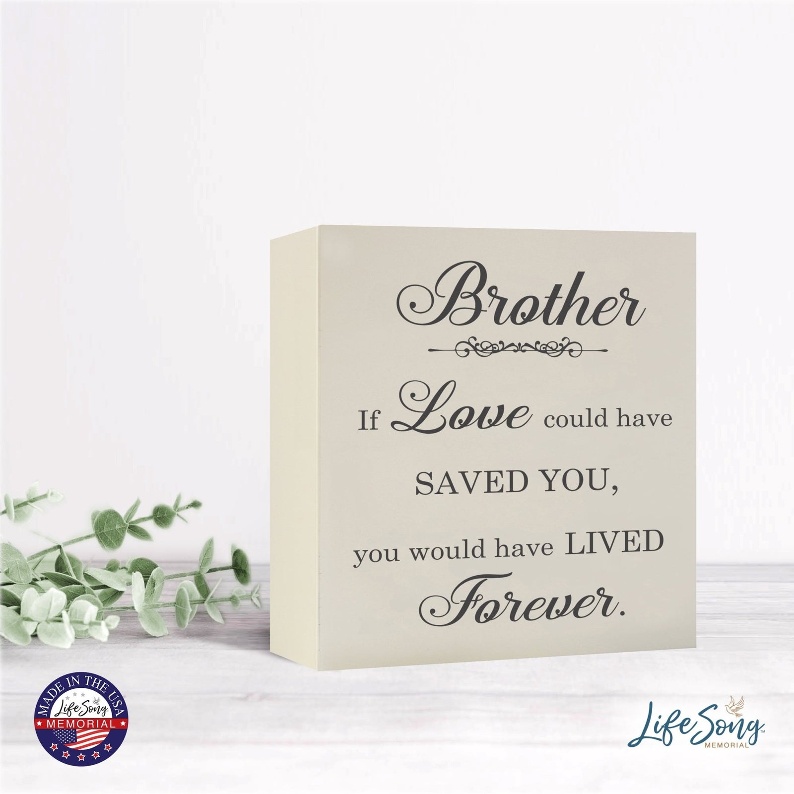 A close-up of the Lifesong Milestones Memorial Shadow Box Urn For Human Ashes, showcasing its heartfelt message, elegantly positioned on a bright, minimalist table adorned with fresh green foliage accents.