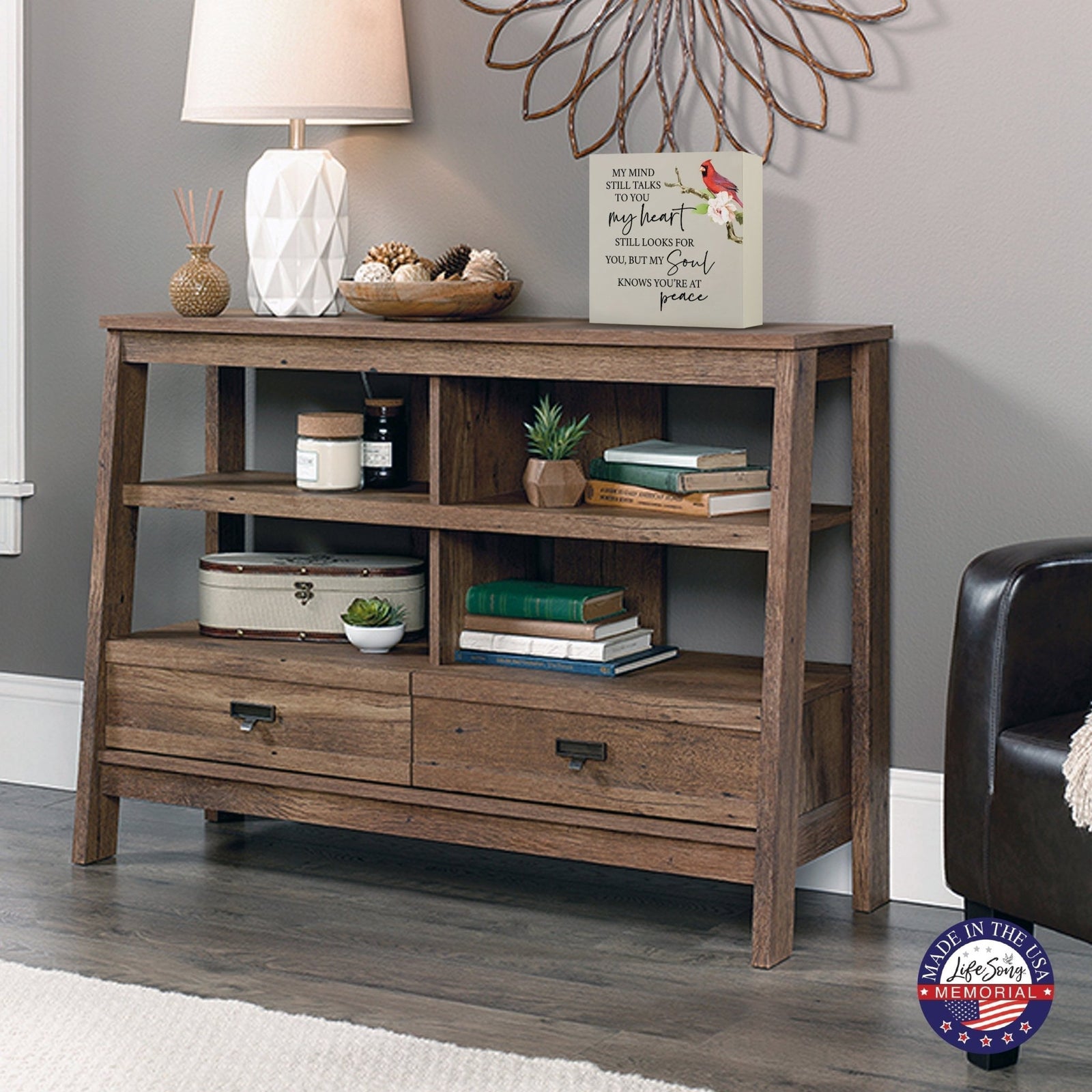 Rustic wooden console table with multiple shelves, decorated with a cream-colored wooden shadow box urn for human ashes. The urn features a red cardinal and floral accents. Surrounded by books, potted plants, and a textured white lamp, this handcrafted adult memorial urn blends seamlessly into a cozy living room setting. Perfect for those seeking wooden shadow box urns for human ashes, offering a beautiful and personalized tribute.