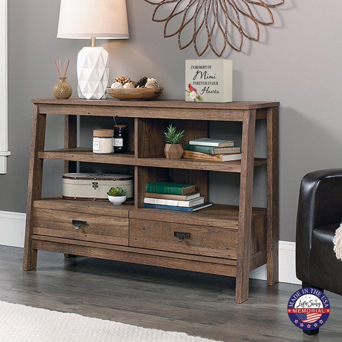 Rustic wooden console table with multiple shelves, decorated with a cream-colored wooden shadow box urn for human ashes. The urn features a red cardinal and floral accents. Surrounded by books, potted plants, and a textured white lamp, this handcrafted adult memorial urn blends seamlessly into a cozy living room setting. Perfect for those seeking wooden shadow box urns for human ashes, offering a beautiful and personalized tribute.