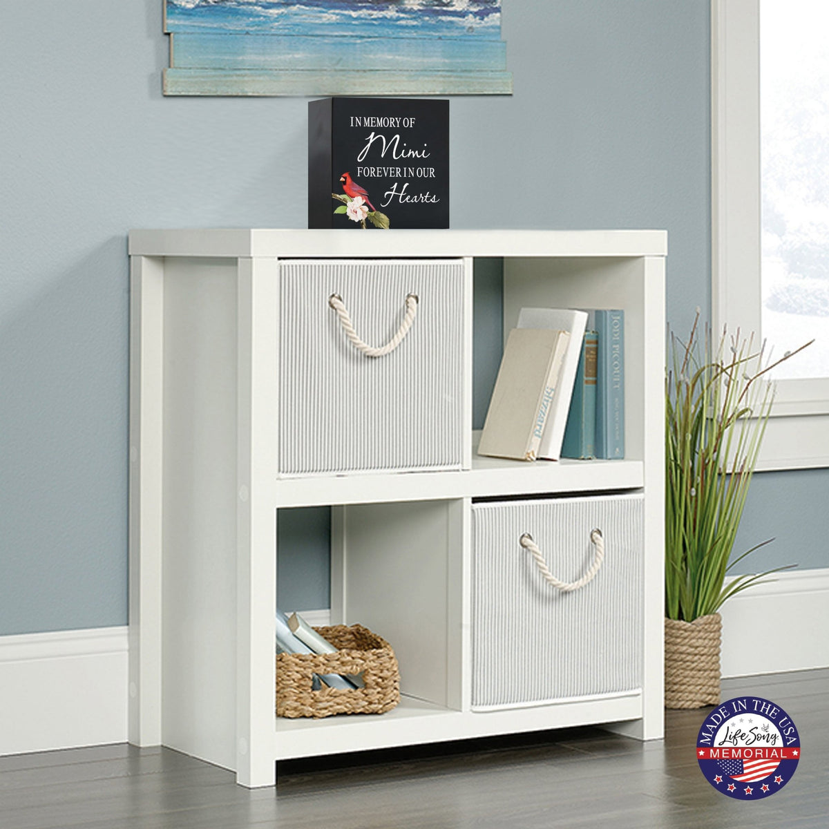 White wooden storage unit topped with a decorative wooden shadow box urn for human ashes. The urn features a red cardinal illustration and floral accents. This adult memorial urn is displayed in a cozy living space with a beach-themed painting above.