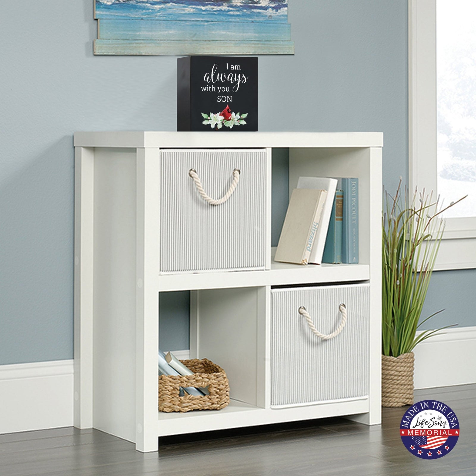 White wooden storage unit topped with a decorative wooden shadow box urn for human ashes. The urn features a red cardinal illustration and floral accents. This adult memorial urn is displayed in a cozy living space with a beach-themed painting above.