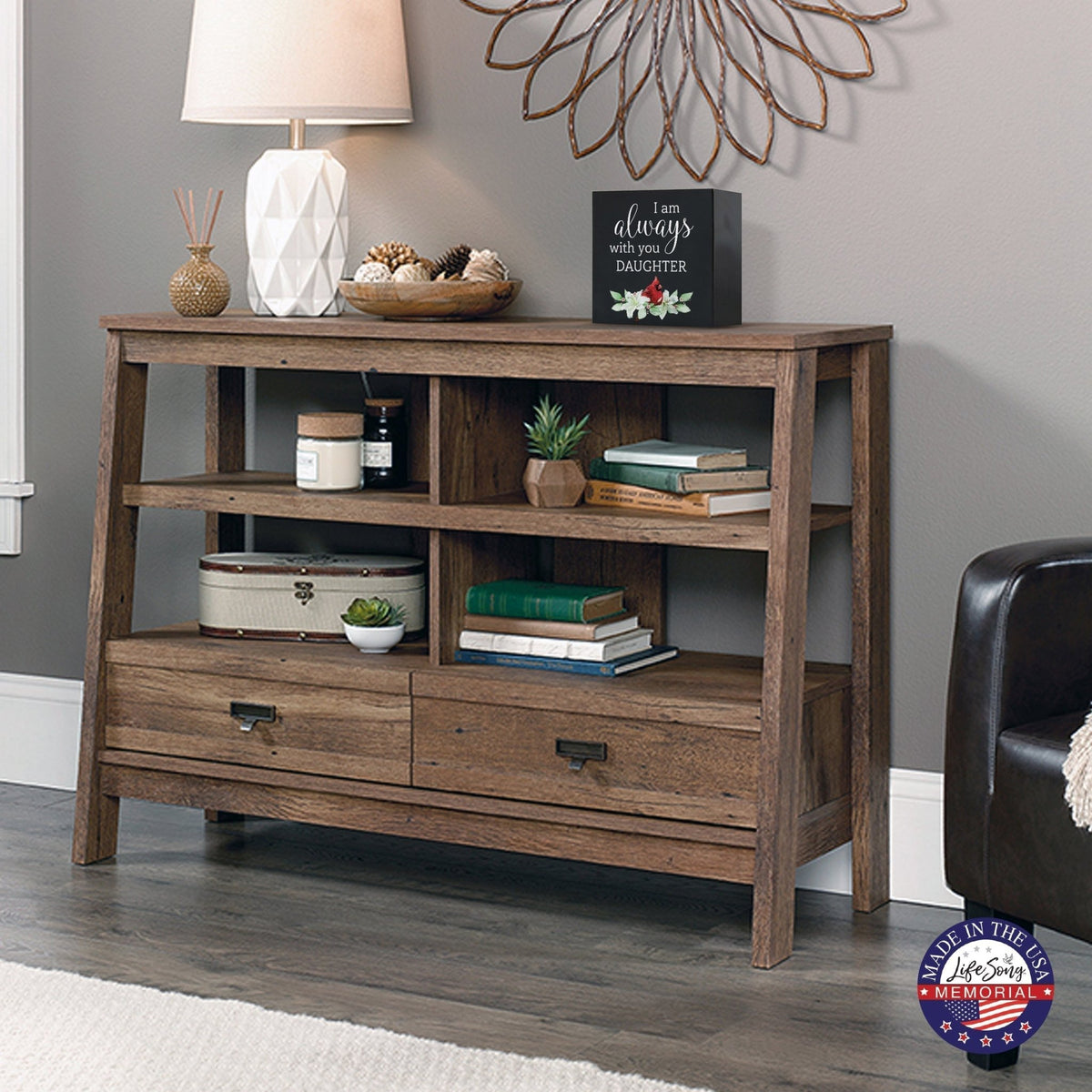 Rustic wooden console table with multiple shelves, decorated with a cream-colored wooden shadow box urn for human ashes. The urn features a red cardinal and floral accents. Surrounded by books, potted plants, and a textured white lamp, this handcrafted adult memorial urn blends seamlessly into a cozy living room setting. Perfect for those seeking wooden shadow box urns for human ashes, offering a beautiful and personalized tribute.