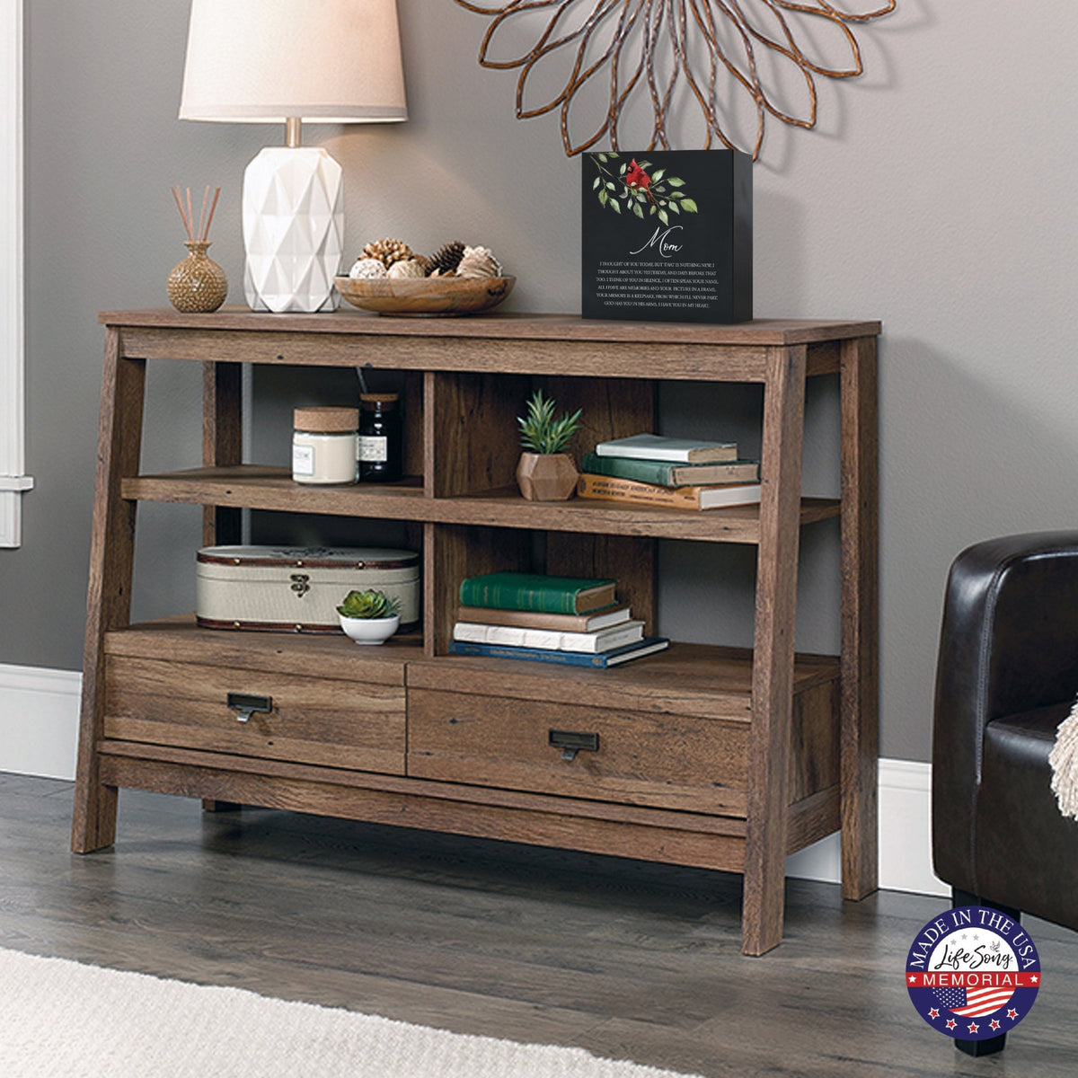 Rustic wooden console table with multiple shelves, decorated with a black-colored wooden shadow box urn for human ashes. The urn features a red cardinal and floral accents. Surrounded by books, potted plants, and a textured white lamp, this handcrafted adult memorial urn blends seamlessly into a cozy living room setting. Perfect for those seeking wooden shadow box urns for human ashes, offering a beautiful and personalized 