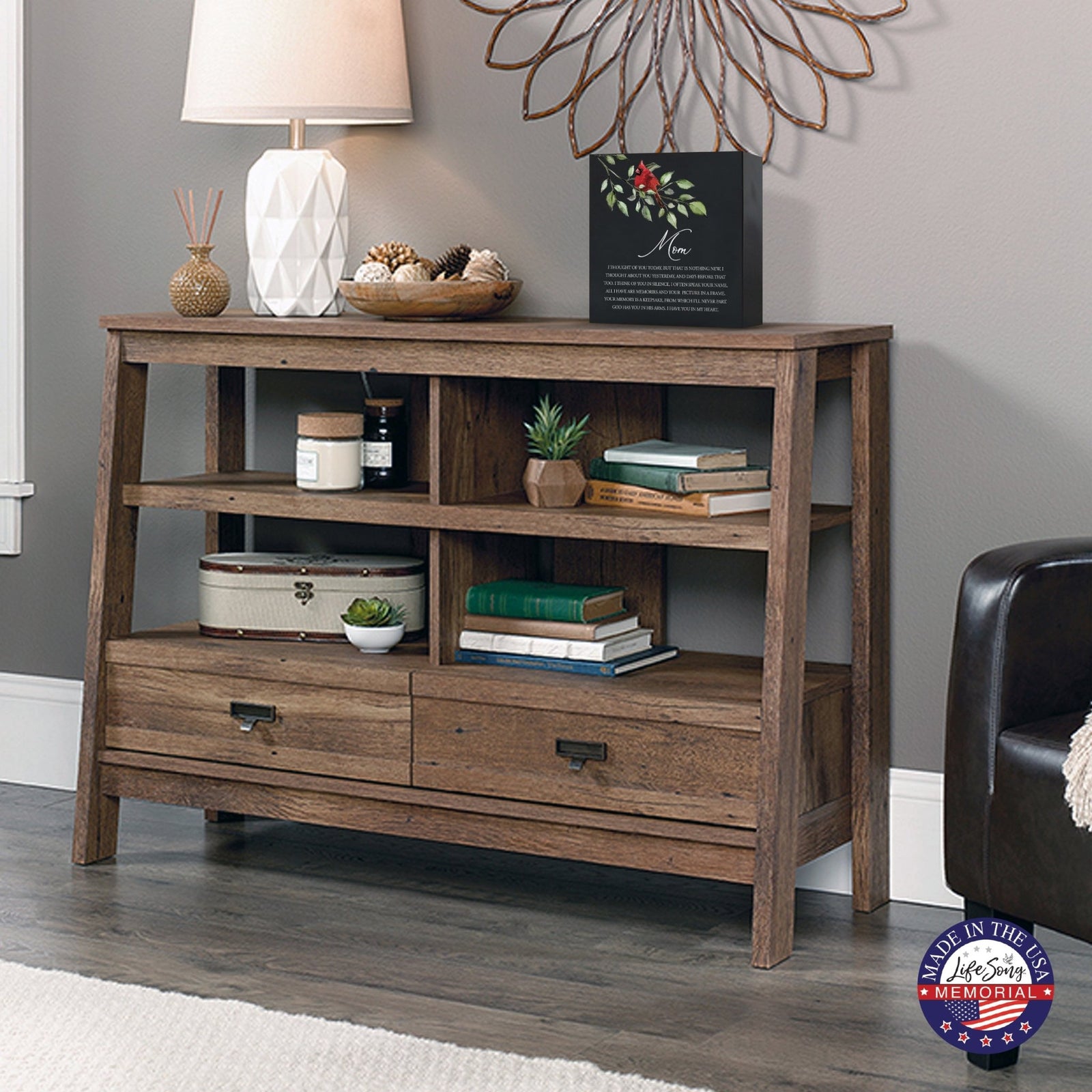 Rustic wooden console table with multiple shelves, decorated with a black-colored wooden shadow box urn for human ashes. The urn features a red cardinal and floral accents. Surrounded by books, potted plants, and a textured white lamp, this handcrafted adult memorial urn blends seamlessly into a cozy living room setting. Perfect for those seeking wooden shadow box urns for human ashes, offering a beautiful and personalized 