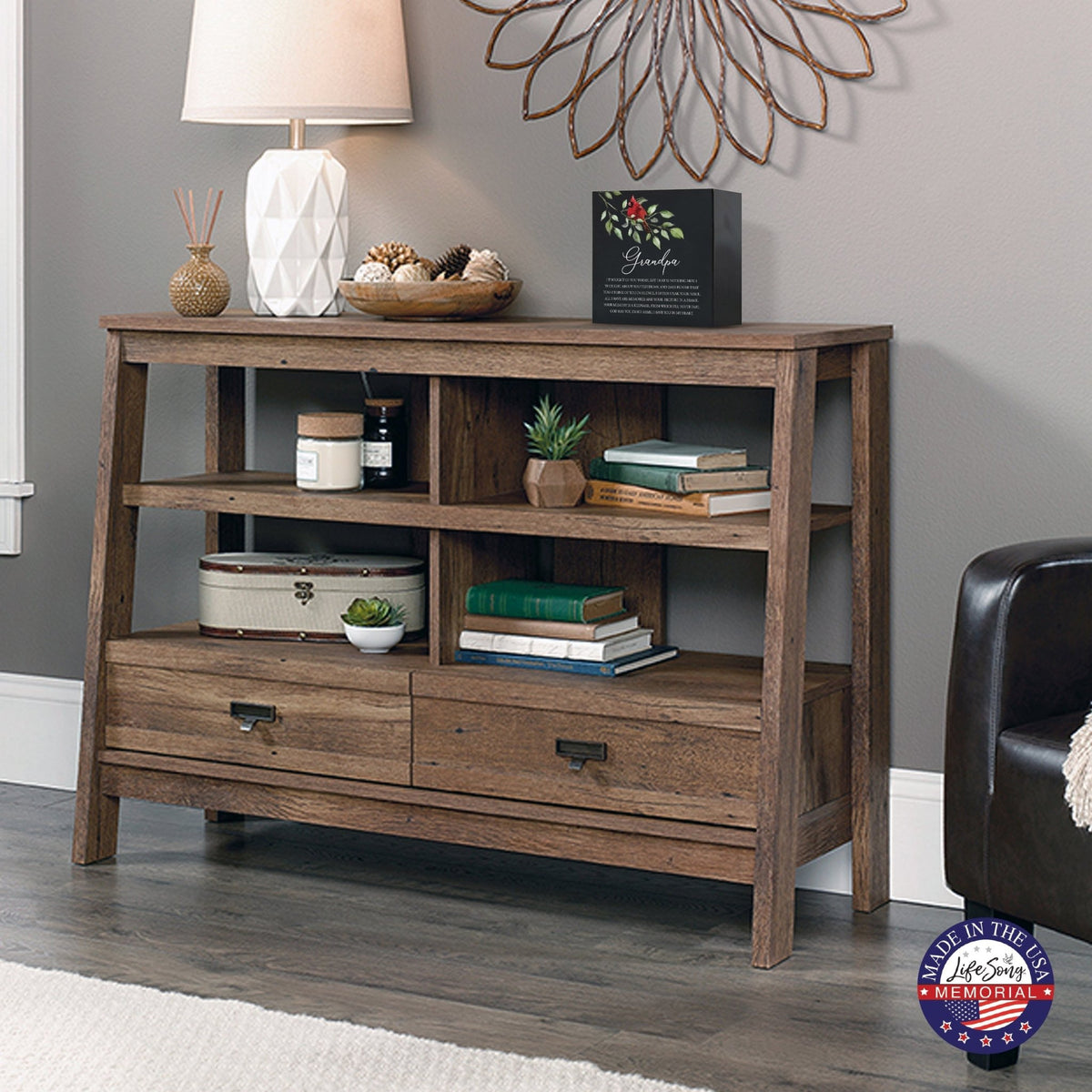 Rustic wooden console table with multiple shelves, decorated with a black-colored wooden shadow box urn for grandpa. The urn features a red cardinal and floral accents. Surrounded by books, potted plants, and a textured white lamp, this handcrafted adult memorial urn blends seamlessly into a cozy living room setting. Perfect for those seeking wooden shadow box urns for human ashes, offering a beautiful and personalized tribute.