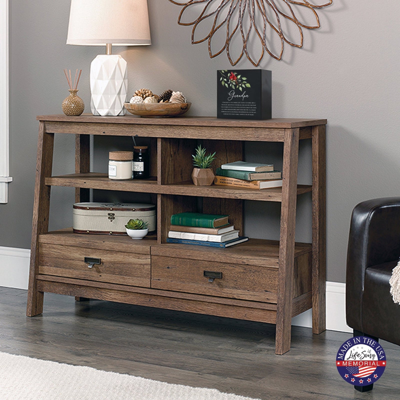 Rustic wooden console table with multiple shelves, decorated with a black-colored wooden shadow box urn for grandpa. The urn features a red cardinal and floral accents. Surrounded by books, potted plants, and a textured white lamp, this handcrafted adult memorial urn blends seamlessly into a cozy living room setting. Perfect for those seeking wooden shadow box urns for human ashes, offering a beautiful and personalized tribute.
