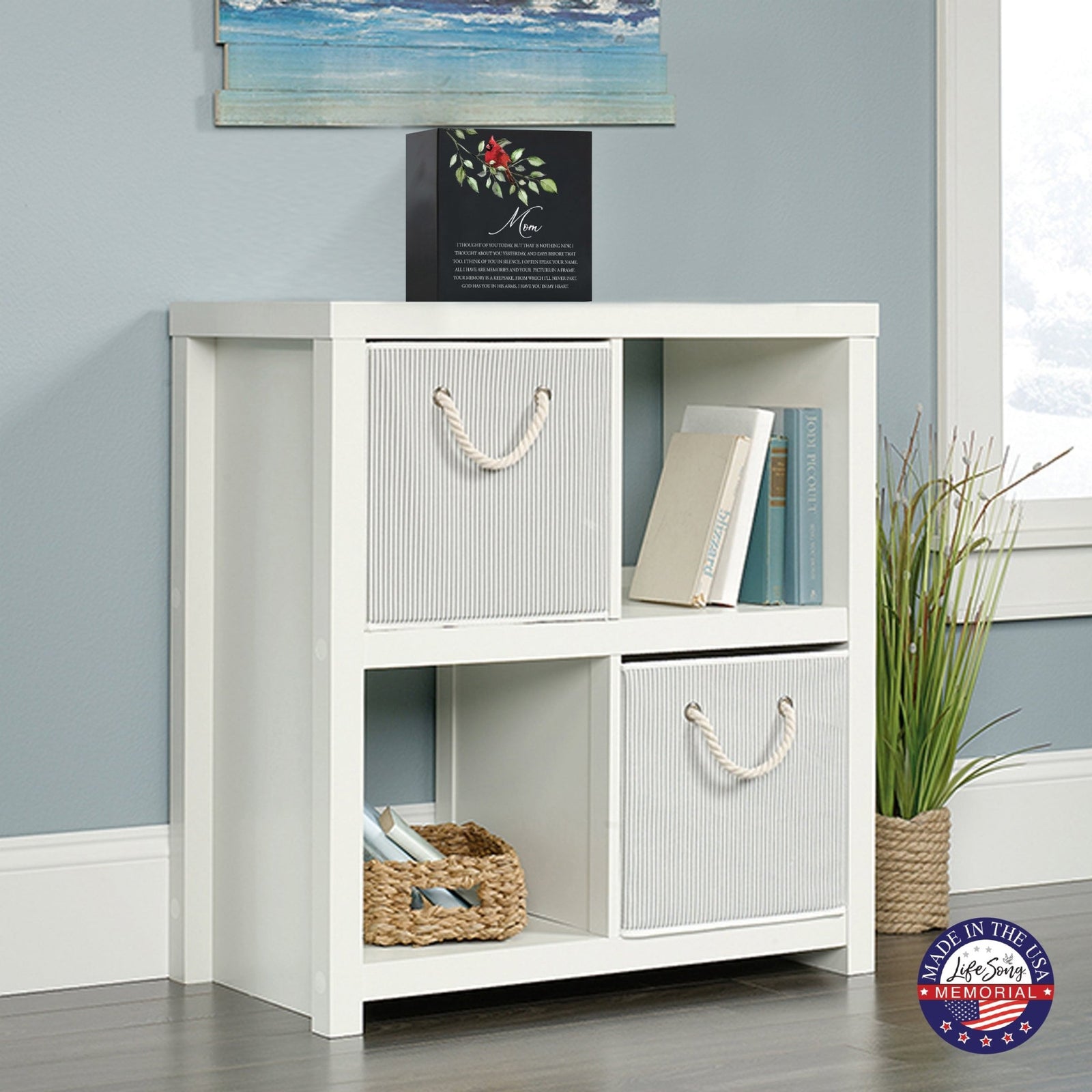 Black wooden storage unit topped with a decorative wooden shadow box urn for mom. The urn features a red cardinal illustration and floral accents. This adult memorial urn is displayed in a cozy living space with a beach-themed painting above.