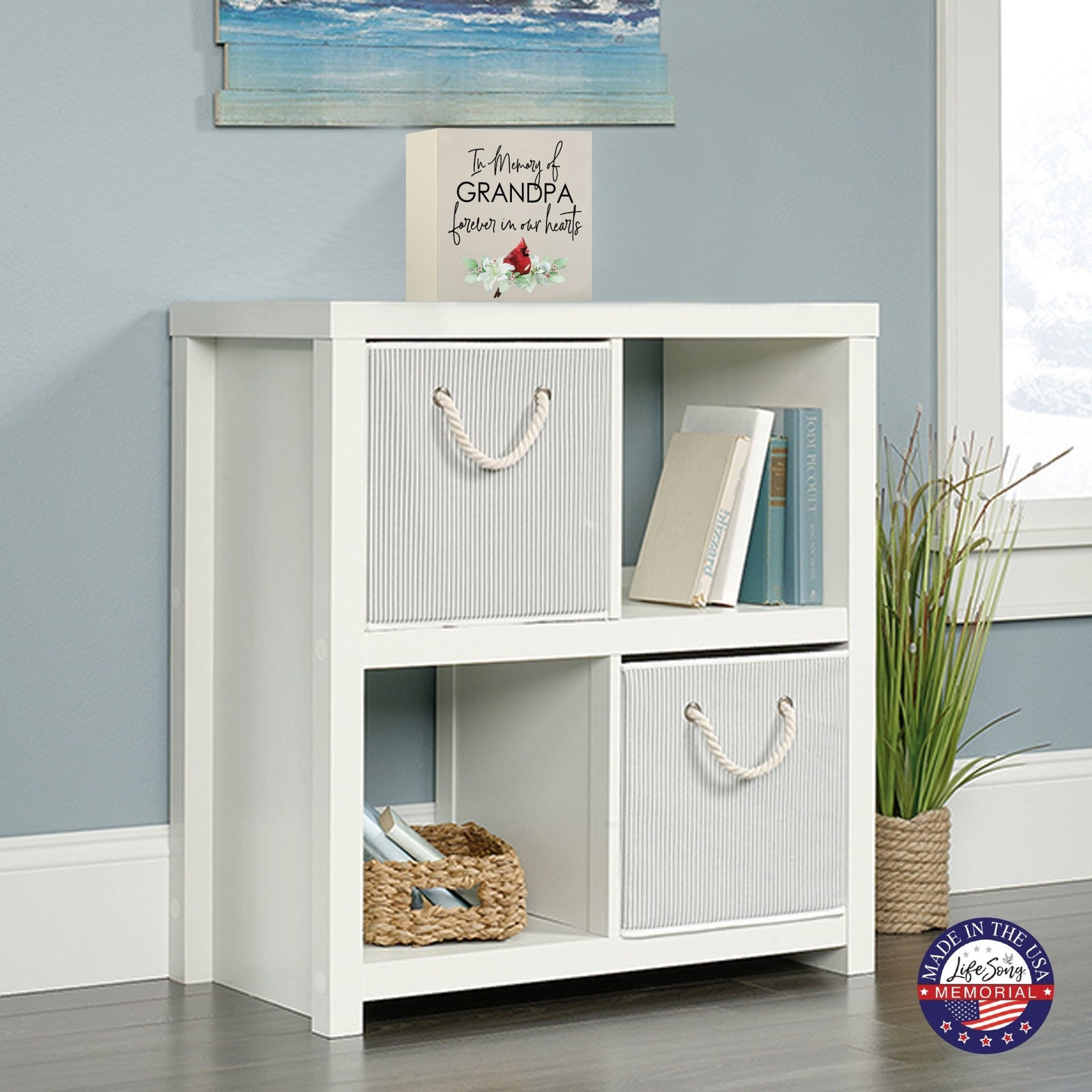 White wooden storage unit topped with a decorative wooden shadow box urn for human ashes. The urn features a red cardinal illustration and floral accents. This adult memorial urn is displayed in a cozy living space with a beach-themed painting above.