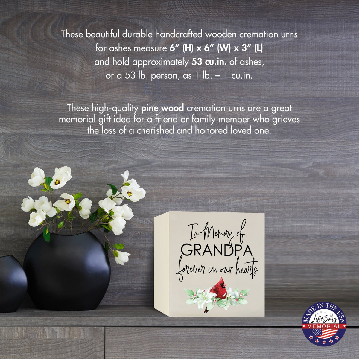 Cream-colored wooden shadow box urn for human ashes, measuring 6" x 6" x 3". The urn is inscribed with "In Memory of Grandpa - Forever in our hearts" alongside a red cardinal and delicate floral design. Displayed on a wooden surface beside modern black vases with white flowers, this handcrafted urn is ideal for honoring adult loved ones.